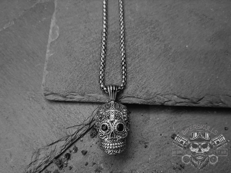 Collana con ciondolo in acciaio dal design accuratamente realizzato raffigurante un Mexican Skull con croce sulla fronte, occhi con cristalli neri e ampi intagli floreali tribali su tutto il cranio.  La giusta collana per attirare l'attenzione desiderata.