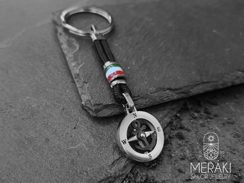 Portachiavi Kai realizzato con cordino nautico nero, charm centrale con bandiera Italiana. A completamento del portachiavi troviamo un charm con ancora girevole nera, rosa dei venti con i punti cardinali a contrasto